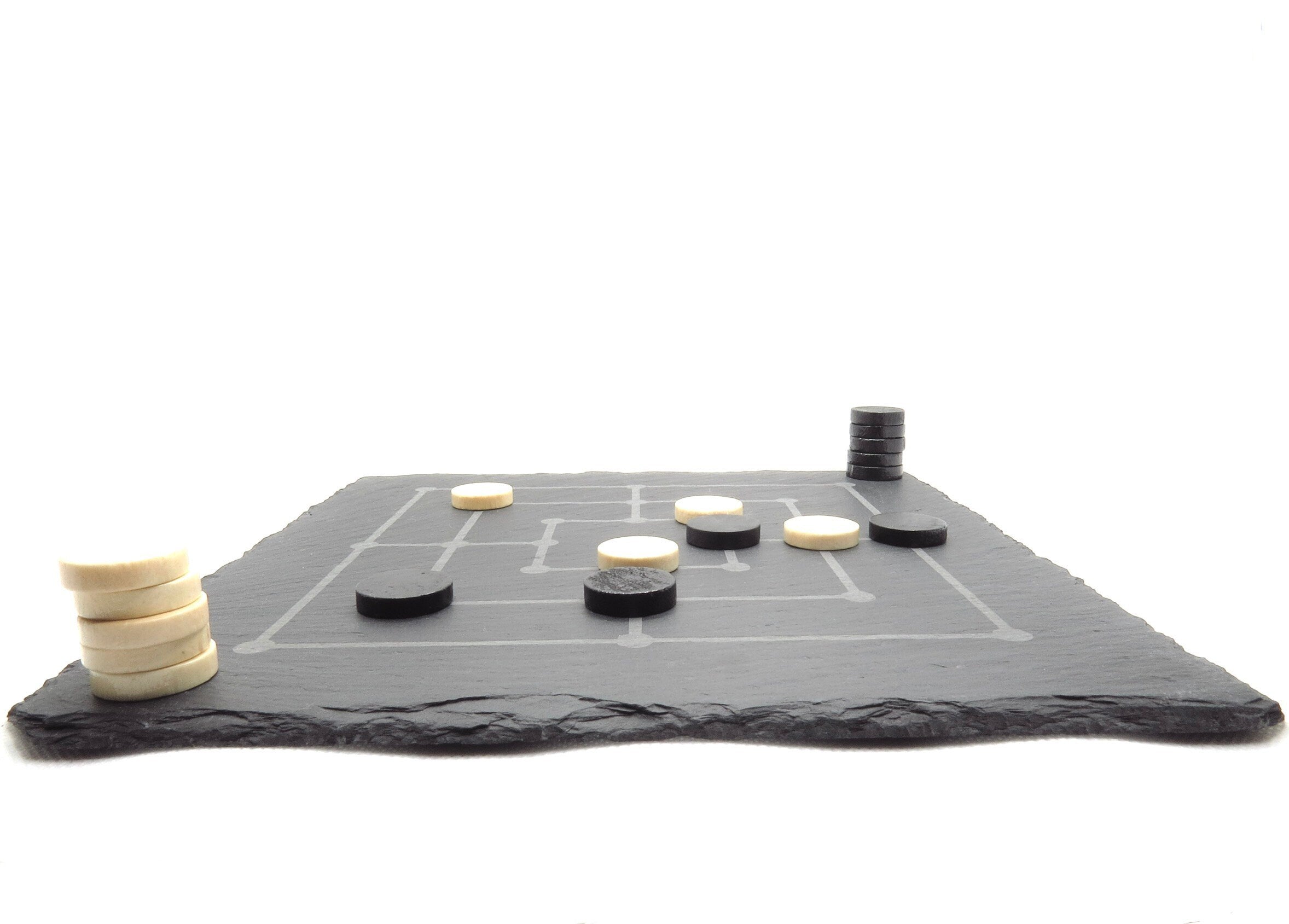 Seitenansicht Mühle-Spiel: Flaches Schieferbrett mit aufliegenden Spielsteinen vor weißem Hintergrund.